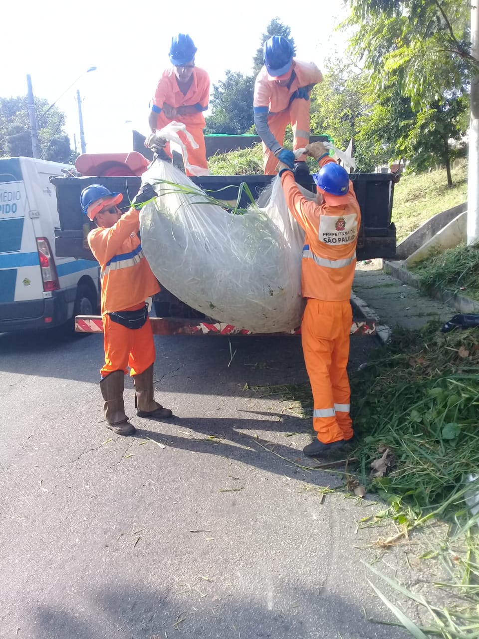 Mato cortado é erguido, enrolado em uma tela de proteção branca, por trabalhadores uniformizados de macacão laranja e capacetes azuis. Dois seguram embaixo e outros dois recebem em cima do caminhão, que já tem uma quantidade de detritos recolhidos. À direita, a praça já com a poda realizada e um pouco de capim cortado sobre a calçada.